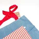 denim blåt materiale med rød og hvid stribet lomme. Hanken er rød med et hvidt mønster.