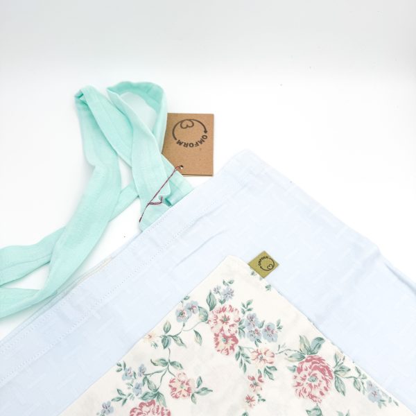 lyseblå materiale med motiv og en beige lomme med blomster. Hanken er turkis.