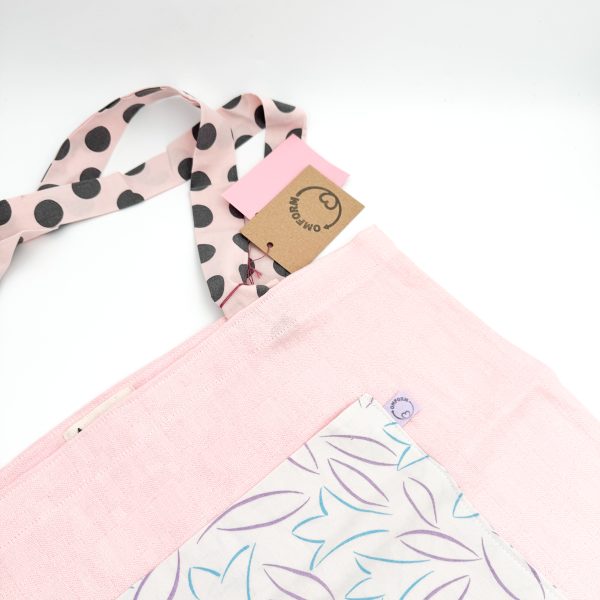 Lyserødt materiale med lidt motiv på. Den har en hvid lomme med flot blomstret mønster i lilla og blå. Hanken er lyserød med store sorte prikker.